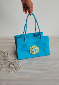 Blue gift bag with a yellow flower design held by a hand on a light wooden surface.